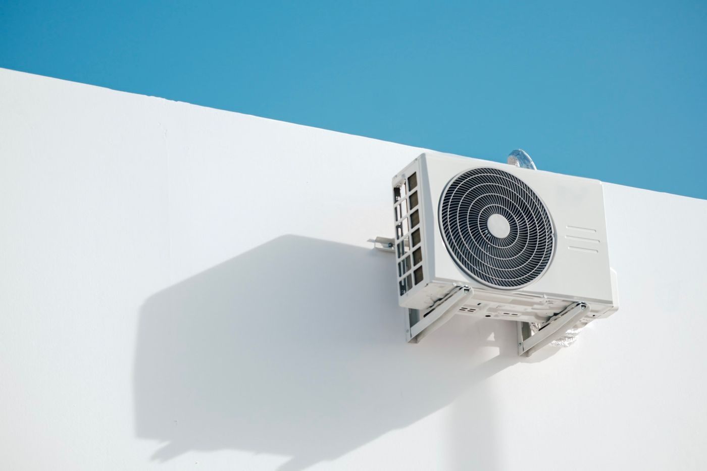 Air conditioning outdoor unit fixed on a wall bracket against a white wall and blue summer sky